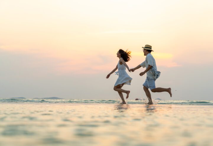 Happy Asian family couple walking and playing in sea water together on tropical beach at summer sunset on beach vacation. Husband and wife relax and enjoy outdoor lifestyle on holiday travel vacation
