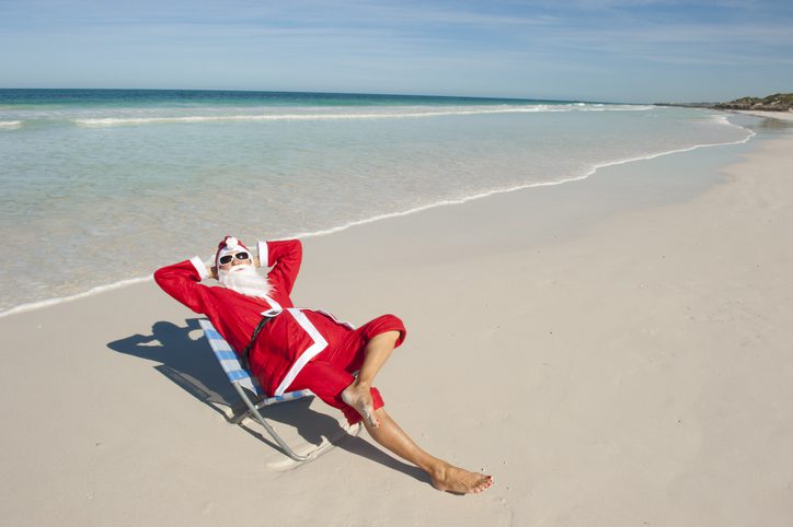 Santa Claus happy relaxed sitting with hands up at beach, having fun and joy off duty at tropical holiday vacation, isolated with ocean and blue sky as background and copy space.
